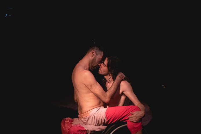 Imagen de fondo negro,  en el centro 2 cuerpos (Javier y Diego), uno de ellos en silla de ruedas, ambos de pantalones rosados, torso y pies desnudos. Están enfrentados uno sentado arriba de otro, abrazados, a un instante de besarse en la boca.