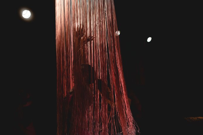 Imagen de fondo negro, de manera protagónica se aprecia una lámpara con tiras finas de color rosado, dentro de la lámpara y debajo de las tiras se ve la sombra de una bailarina con brazos extendidos hacia arriba.